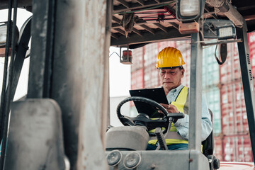 Male worker wearing a helmet inspects the order of the container he sits on a forklift and takes notes.