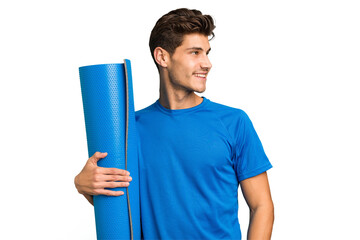 Young caucasian sport man holding a mat isolated looks aside smiling, cheerful and pleasant.