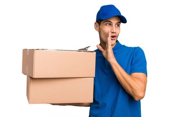 Young delivery caucasian man holding boxes isolated is saying a secret hot braking news and looking...