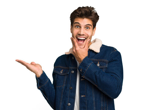 Young caucasian handsome man isolated holds copy space on a palm, keep hand over cheek. Amazed and delighted.