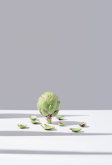 Flor de alcachofa cruda en pie con hojas que caen sobre una mesa blanca y fondo gris	