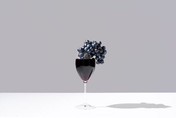 Un racimo de uvas negras dentro de una copa de vino tinto goteando sobre fondo gris