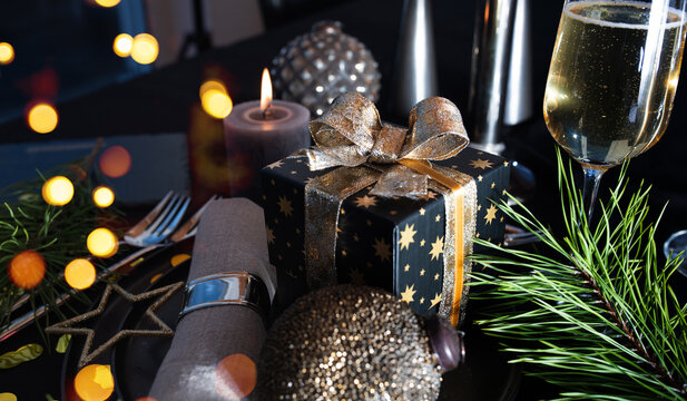 Atmospheric table decoration for a christmas or new year dinner. Place setting in modern style. Champagne, gift box, festive branch decoration with candle and golden bokeh on black tablecloth.