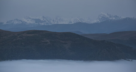 Low clouds in the mountains filled the entire valley and rocky peaks and hills with trees stick out above the clouds, mountains with snow in the background, fog between the mountains on an autumn