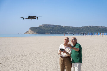 coppia di anziani si diverte con un drone che pilotano in volo in una spiaggia con il cielo blu - obrazy, fototapety, plakaty