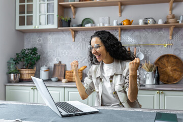 Excited and happy hispanic woman celebrating winning the lottery by raising her hands. Sitting at home in the kitchen with a laptop, receiving a new job opportunity, received good news.