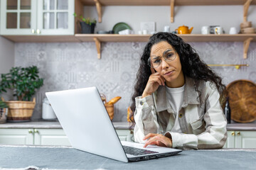 Tired young beautiful Latin American woman sitting at home at the laptop, bored from work. He holds his head with his hand and looks at the camera.