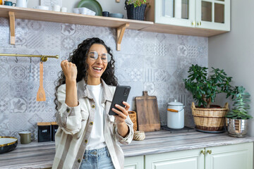 Happy beautiful young hispanic woman messing around in the kitchen at home with her phone, playing...