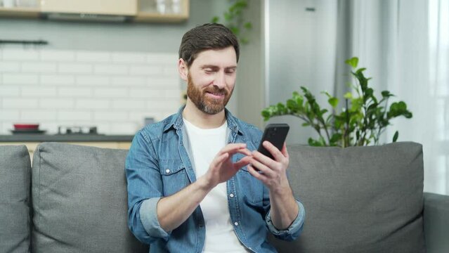 Single bearded man swiping through women profiles on dating app and like or dislike photos on smartphone in the living room Young guy looking for partner on social platform and waiting for a match