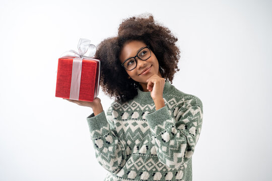 Portrait of young attractive african american woman with curly hair and glasses presenting gift box in studio on white background.