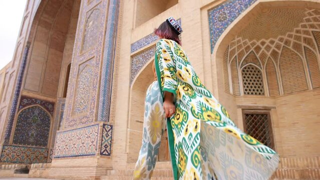 An oriental girl in a traditional Uzbek dress walks past an old mosque. Landmark of Bukhara. Back view. Eastern madrasah.