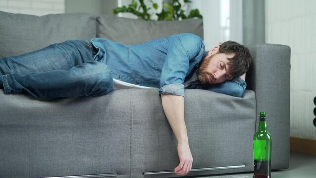 Portrait of drunk bearded man sleeping on the couch at home with a bottle next to him and has a severe hangover after big spree Alcoholism addiction and bad habits concept
