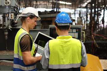 Technician engineer in protective uniform with hardhat standing and teaching apprentices or colleague worker to use computerized machine control at heavy industry manufacturing factory