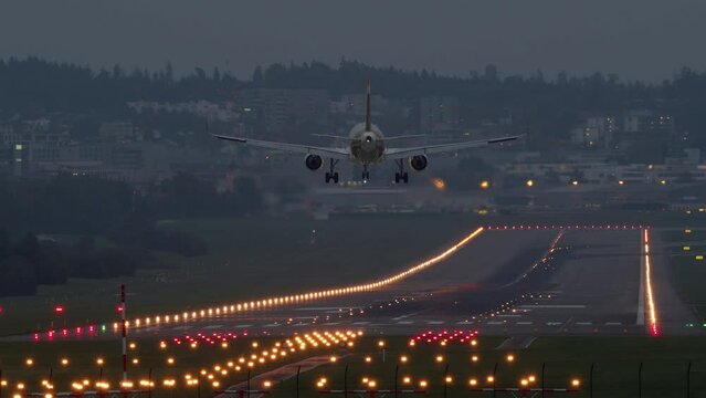 A passenger airplane with side lights on, lands at dusk on a brightly lit runway - Long shot