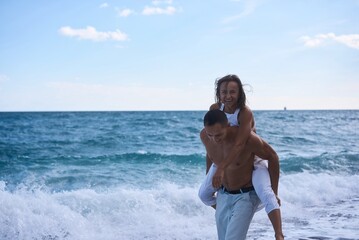Happy couple in love on beach summer vacations. Joyful girl piggybacking on young boyfriend having fun.
