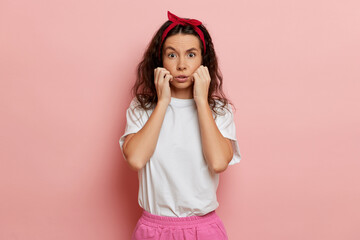 Woman shows scare emotion, stands isolated over pink, keeps arms close to her face