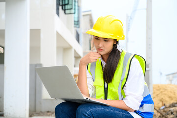 Young attractive construction woman smiling with yellow helmet working with laptop, sitting at building construction site. Home building project.