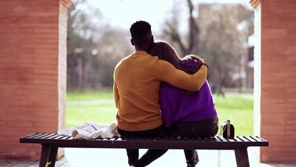Friend embracing girlfriend, black man putting arm around girl sitting at park bench