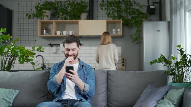 lustful man texting other female lovers sitting on the couch while wife does housework and noticed it Perplexed bearded man feels confused while his wife screaming hitting him Cheating lie concept 