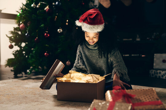 Christmas, gift and girl opening box at night in home on holiday. Xmas spirit, happy and excited child or kid with smile, looking at glowing presents or gifts and enjoying December holidays in house.
