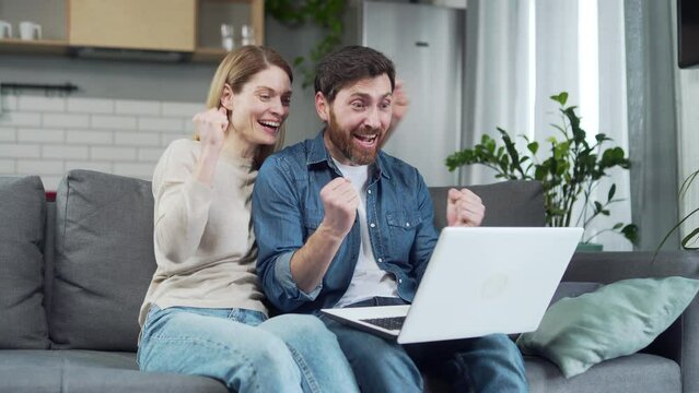 Amazed lucky couple extremely happy after read message in laptop at home Excited young family looking at screen and pleased rejoices received great news on computer Moment of success triumph