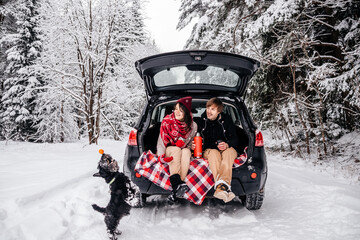 a young couple, a guy and a girl, are sitting with a dog in the trunk of a car in the winter in the forest