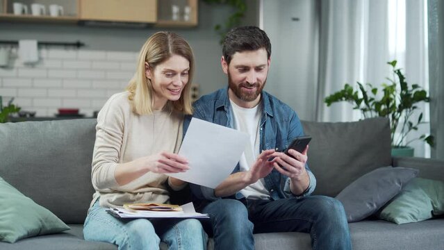 Positive young family do paper work paying utility bills Bearded smiling husband making payment transaction using online application on phone in living room Finance management and easy pay concept