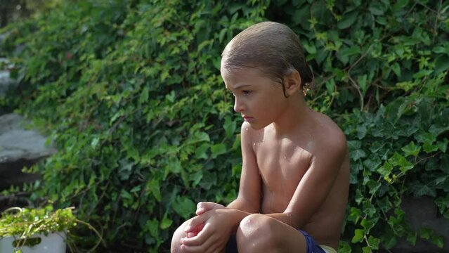 One pensive young boy sitting outside after bathing drying in the sun. Thoughtful wet child resting after swimming wearing swimwear shorts. Contemplative emotion
