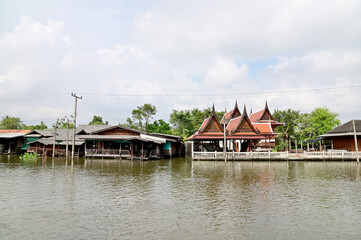 Fototapeta premium BANGKOK, THAILAND - NOVEMBER 20, 2022 : Wooden houses, Thai houses and buildings along the Chao Phraya River with white clouds and blue sky background, Town Ayutthaya Waterfront, Thailand.