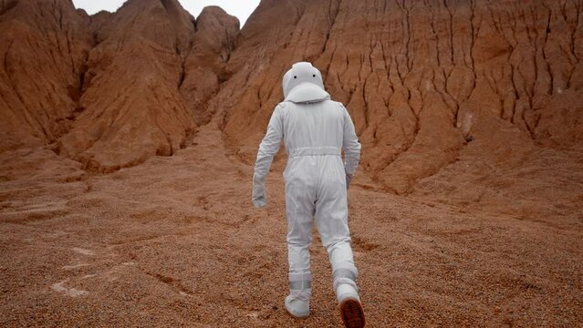 A view from behind an astronaut in a white suit and helmet walking towards the red hills. Astronaut in the crater on the unknown planet goes to explore the surface.