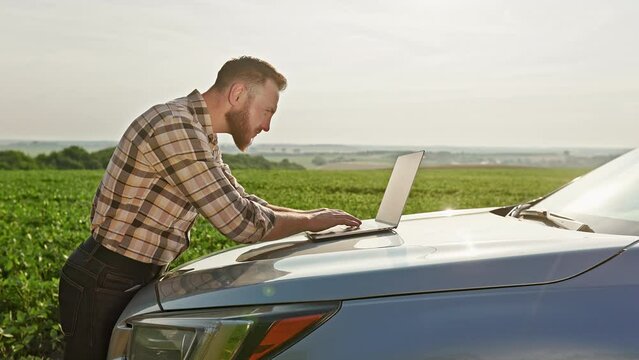 A farmer working on a laptop on the hood of a car standing in the middle of a field. He turned and looked at the field. He covers himself from the sun with his hand.