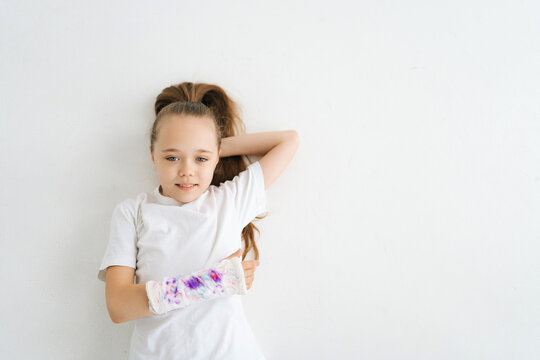 High-angle view of positive sweet little girl with broken hand wrapped in plaster bandage with colorful draw lying on white floor in studio looking at camera. Concept of child insurance and healthcare