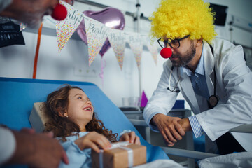 Happy doctor with clown red noses celebrating birthday with little girl in hospital room.