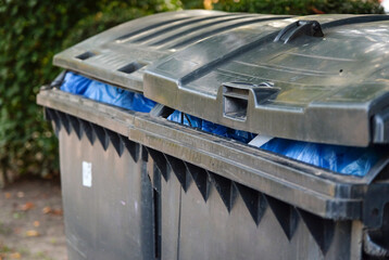 Seitliche Nahaufnahme von zwei mit blauen Müllsäcken randvoll gefüllten Mülltonnen –...