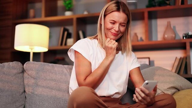 Charming emotional woman admires family kid photos or cute internet pictures on smartphone gadget sitting on sofa in the living room.  