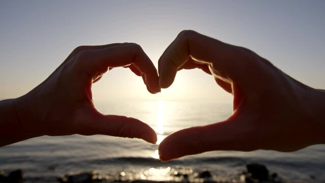 Male and female hands making heart shape against sunset and sea.