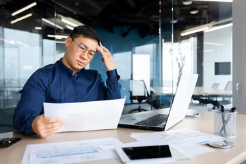 Desperate businessman looks and reads financial reports and documents, Asian boss works with documents inside office, confused and depressed.