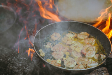 A large pan of shrimp in  is cooked above an open flame set in an outdoor