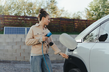 Young woman holding power supply cable from her electric car, prepared for charging it in home,...