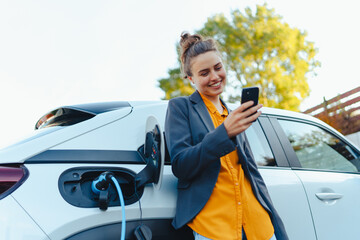 Young woman with smartphone waiting while her electric car charging in home charging station,...