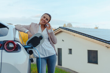 Young woman with smartphone waiting while her electric car charging in home charging station,...