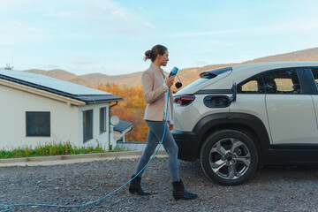 Young woman holding power supply cable from her electric car, prepared for charging it in home,...