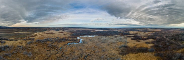 Drone view of a dry looking marsh with a winding stream under a sky filled with white to gray clouds.
