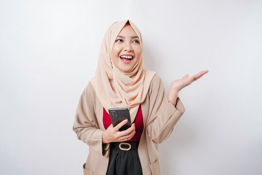 Excited Asian woman wearing hijab pointing at the copy space beside her while holding her phone, isolated by white background