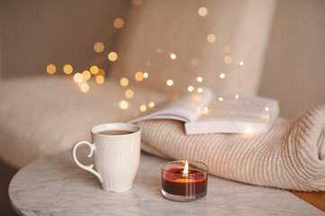 Fototapeta na wymiar Coffee pot with cup of tea burning scented candle on marble table over Christmas lights in bedroom. Cozy home atmosphere.