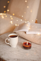 Cup of black tea with paper open book and burning scented candle on marble table over Christmas lights in bedroom. Cozy home atmosphere.