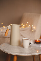 Coffee pot with cup of tea burning scented candle on marble table over Christmas lights in bedroom. Cozy home atmosphere.