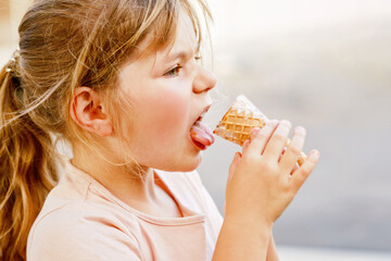 Little adorable preschool girl eating ice cream in waffle cone on sunny summer day. Happy toddler...