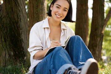 Portrait of happy asian girl sitting in park and writing in her diary. Young woman doing homework on fresh air, sitting near tree and smiling