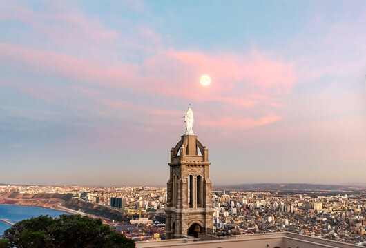 Aerial view from Santa Cruz Chapel tower and the virgin Mary statue back from behind looking at a full moon and the city of Oran with a colorful cloudy sky blue hour sunset twilight and water seaport